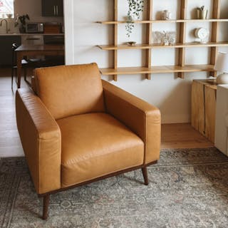 Tan leather armchair with walnut legs in a living room with wood shelving on the wall.