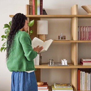 Person in green sweater reading a book in front of the Torme 48" Bookcase, Tall in White Oak finish, styled with plants in a sunny room