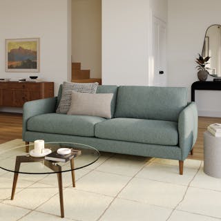 A green fabric sofa with walnut legs in a living room with a glass coffee table.