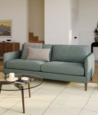 A green fabric sofa with walnut legs in a living room with a glass coffee table.