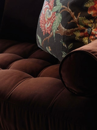 Close-up of a tufted brown velvet sofa with a floral-patterned pillow.