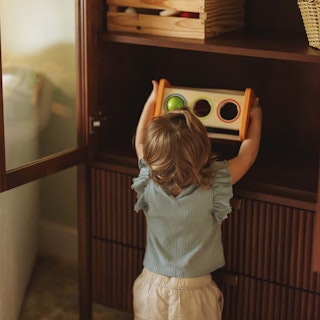 A small child putting away a toy into the Liv 35.25" Glass Cabinet in Walnut